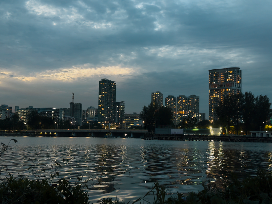 City skyline at twilight, reflecting lights on calm water under a cloudy sky.