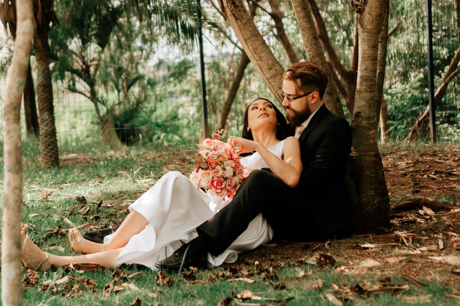 Bride and groom sitting under a tree with a bouquet, enjoying a serene moment together.