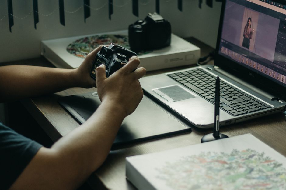 Photographer using a laptop and stylus for photo editing, holding a camera in an indoor setup.