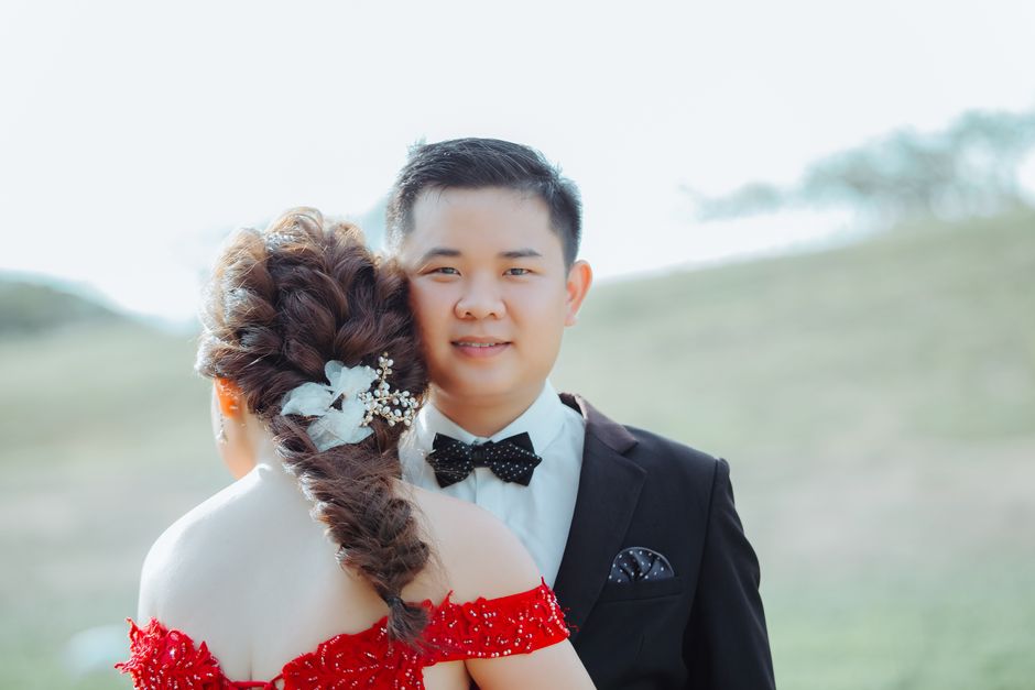 Anonymous woman in bright stylish wear with creative hairstyle decorated with flower embracing content ethnic boyfriend in suit looking at camera on wedding day near grass hillside under sky