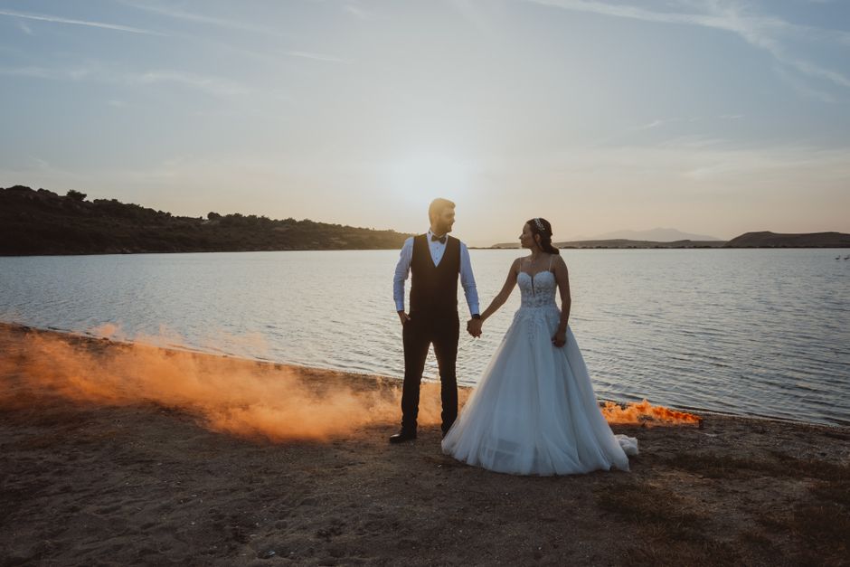 A bride and groom holding hands near a scenic lake with vibrant smoke during sunset.